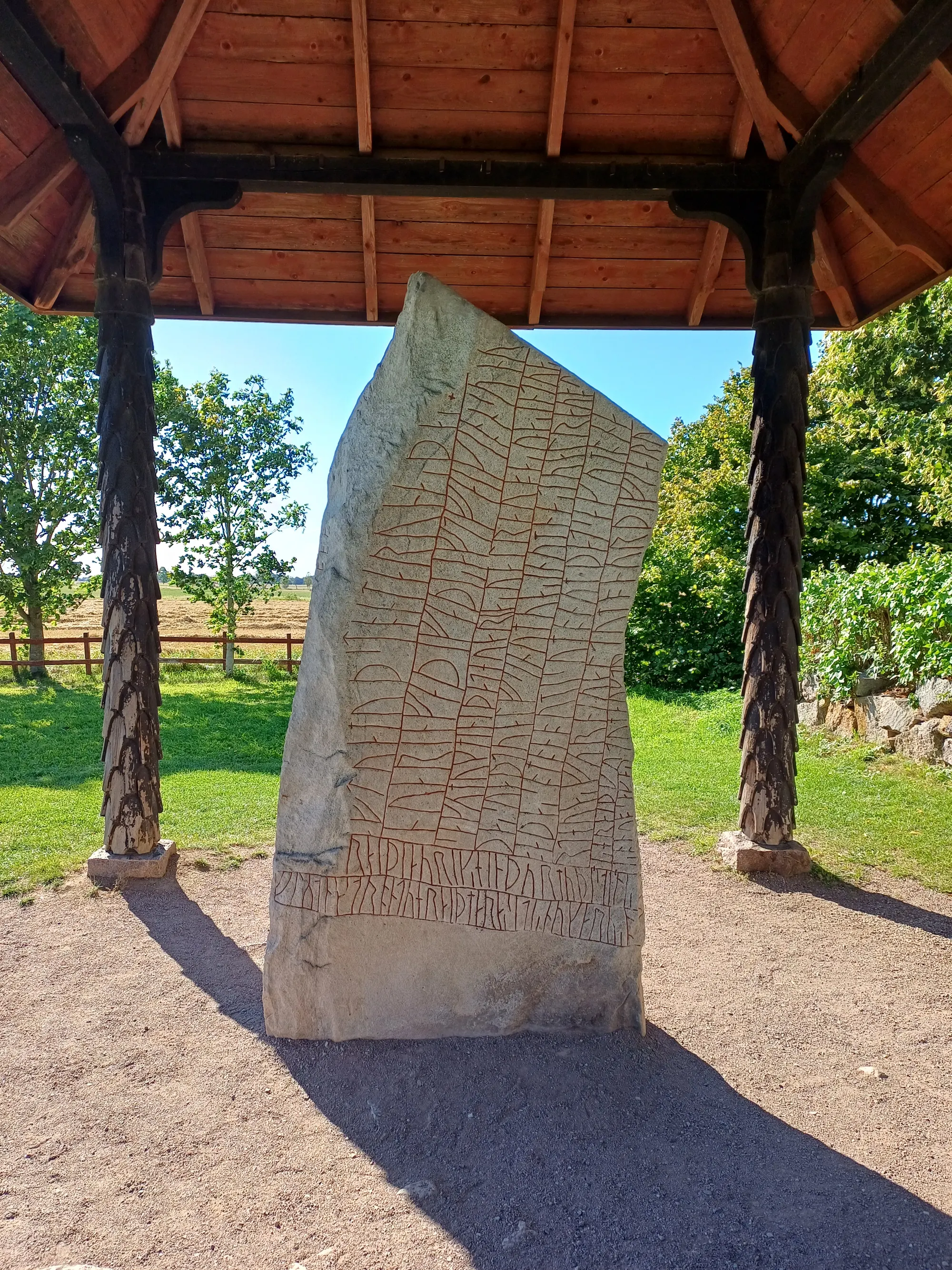 Tall carved stone covered in lines of runes, standing under a wooden shelter roof, with grass and trees behind
