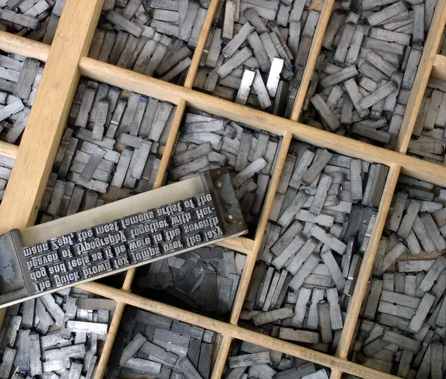 Metal type sorts heaped in a wooden type case, a composing stick of set, mirror-image type across them; photo, 2004
